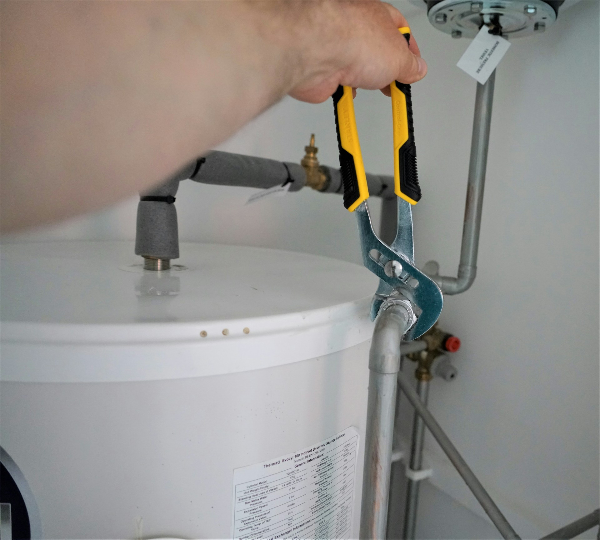 Plumber using yellow-handled pliers to repair pipe connection on white water heater tank with specification label visible
