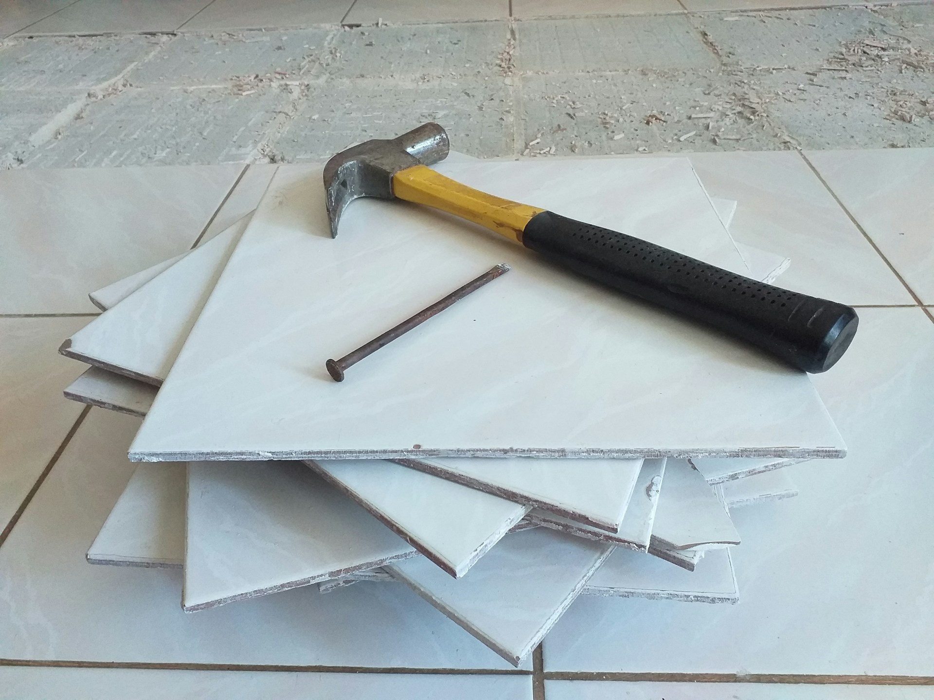 Tiling hammer with yellow handle and black grip alongside broken white ceramic tiles and metal tile spacers on tiled floor
