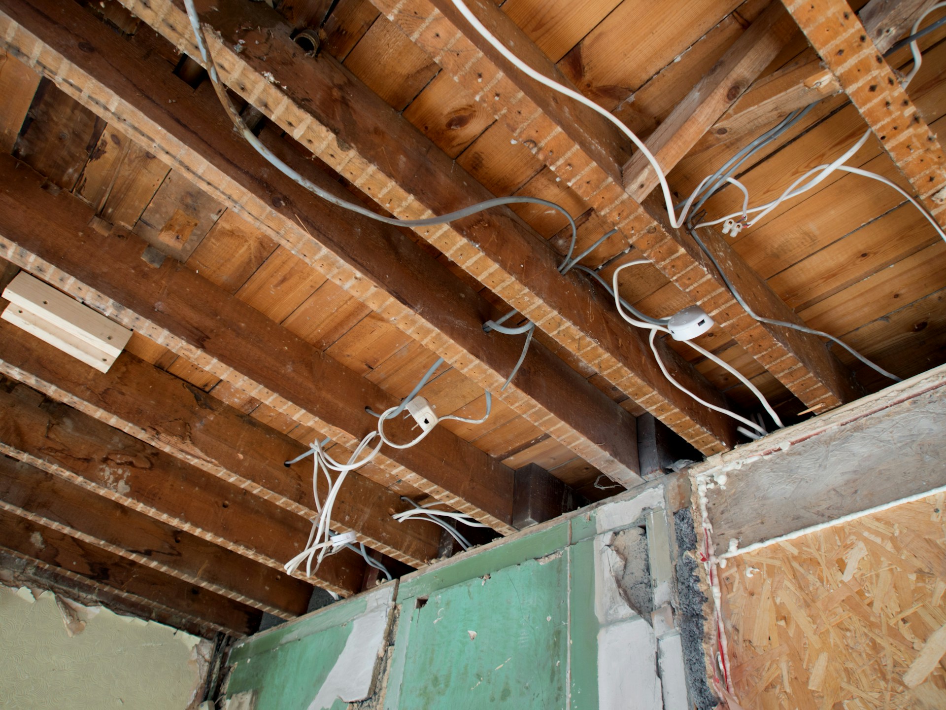Exposed wooden ceiling joists showing new white electrical cables and junction boxes during house rewiring first fix stage with green plasterboard visible