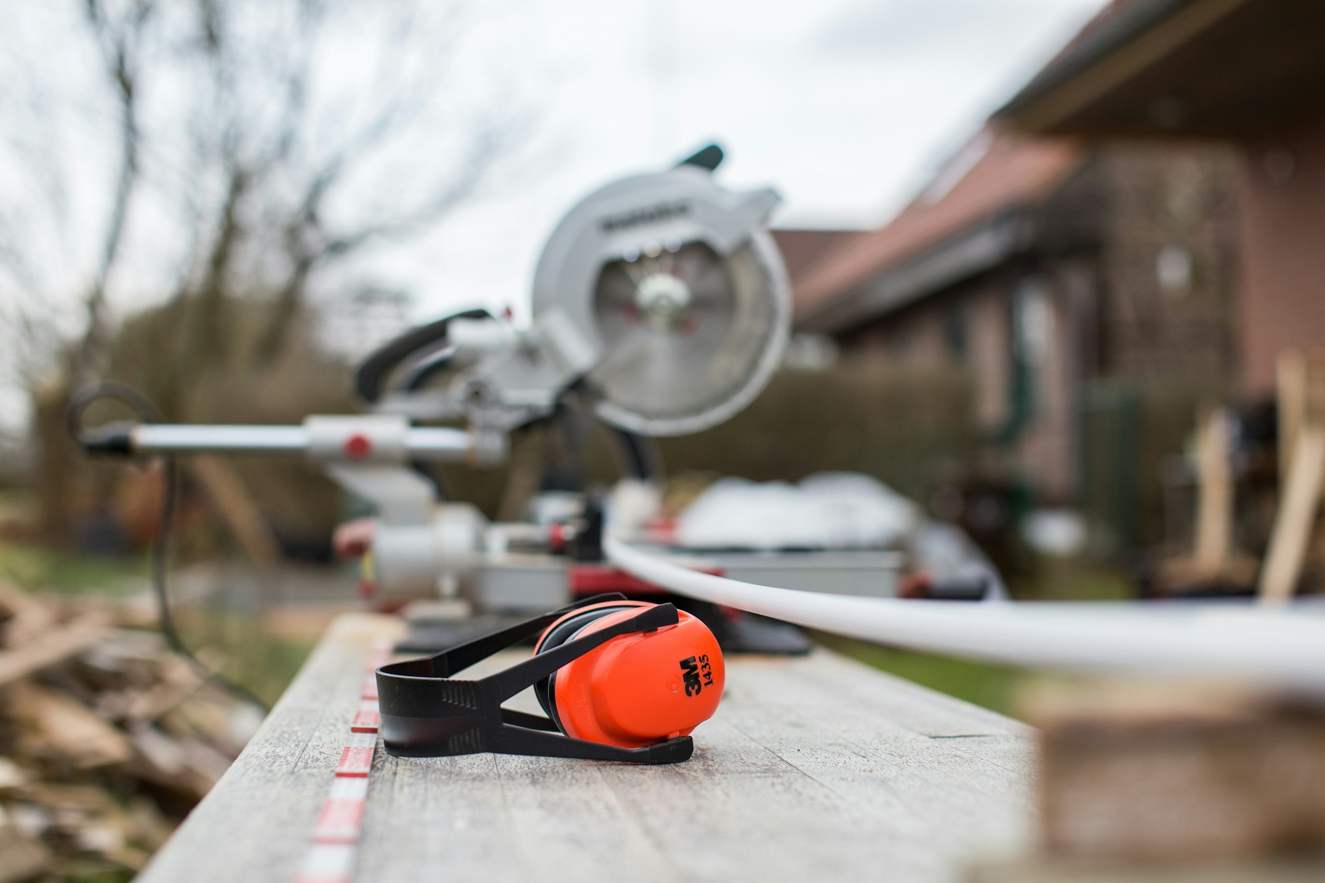 Orange 3M ear defenders on wooden plank with circular saw in background at UK carpentry workshop